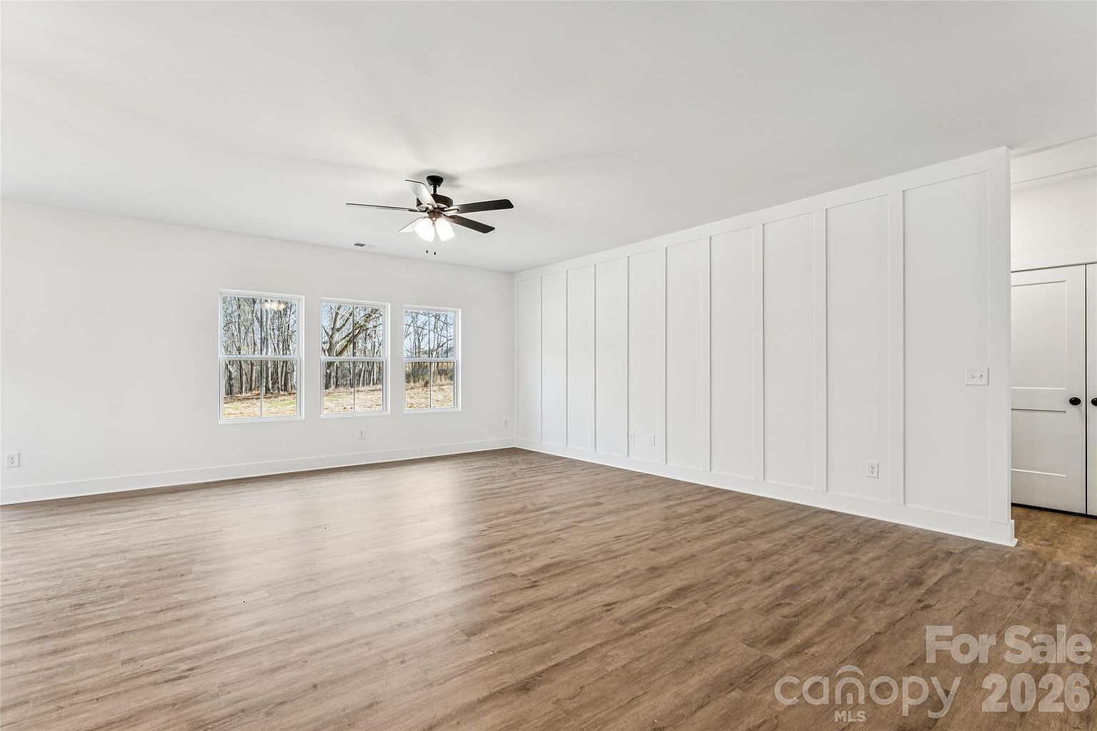 Bonus room or additional bedroom with white board-and-batten accent wall, ceiling fan, and three windows with a wooded view